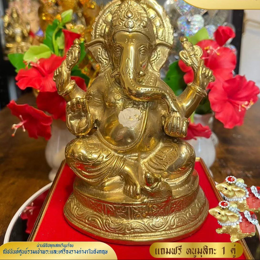A golden brass Ganesha statue seated on a red cushion, displayed on a wooden table with red flowers and other decorative items in the background. Thai text overlays are present in the corners.