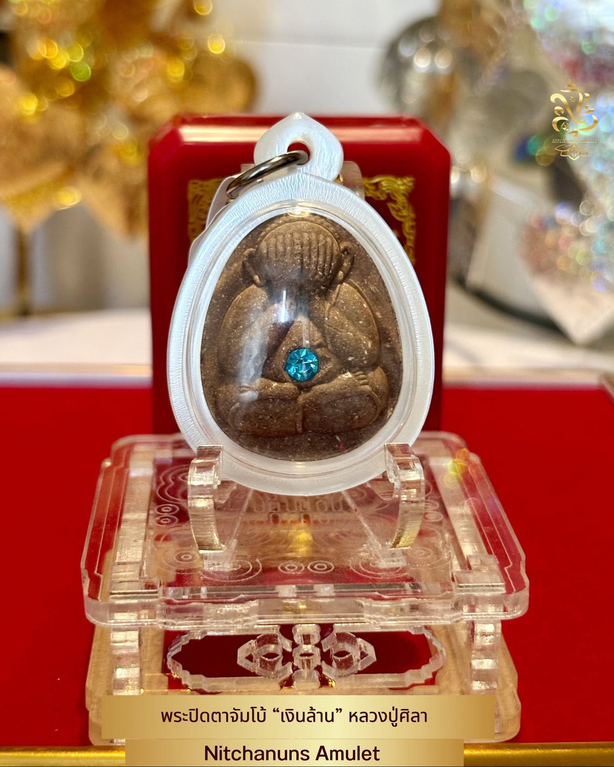 A Nitchanuns Amulet displayed on a clear acrylic stand, featuring a brown seated Buddha figure inside a white oval case with a small blue gemstone at the center, set against a red background with blurred festive lights.
