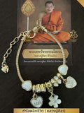 A gold charm bracelet with multiple heart, flower, and cylindrical charms, displayed in front of a printed card featuring a monk. The bracelet has white inlaid accents on several charms.