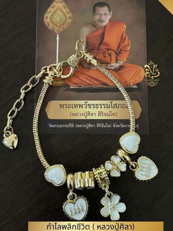 A gold charm bracelet with multiple heart, flower, and cylindrical charms, displayed in front of a printed card featuring a monk. The bracelet has white inlaid accents on several charms.