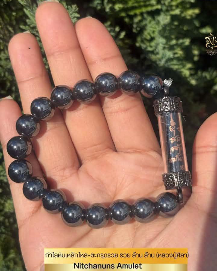A beaded amulet bracelet displayed on an open hand, featuring dark polished round beads and a clear cylindrical amulet with a black and gold calligraphic insert. The background is blurred green foliage.